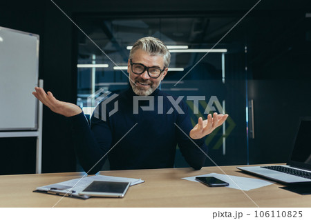 Portrait of an older male businessman sitting in the office smiling and talking on a video call, conducting a webinar, an online business meeting. Portrait of an older male businessman sitting in the office smiling and talking on a video call, conducting a webinar, an online business meeting. 106110825