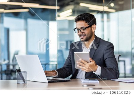 Young successful hispanic businessman at workplace using app on tablet computer, smiling man inside office building, working with laptop, wearing business suit, happy with achievement. Young successful hispanic businessman at workplace using app on tablet computer, smiling man inside office building, working with laptop, wearing business suit, happy with achievement. 106110829