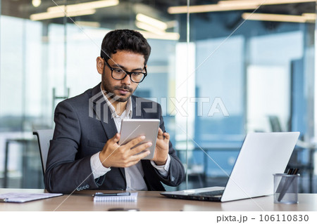 Serious mature concentrated arab businessman working inside office at workplace, man in business suit holding tablet computer, boss using app and laptop thinking. Serious mature concentrated arab businessman working inside office at workplace, man in business suit holding tablet computer, boss using app and laptop thinking. 106110830