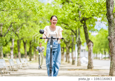 緑が美しい並木道を電動キックボードで爽快に走る若くてかわいい女性｜電動キックボードイメージ 106116863