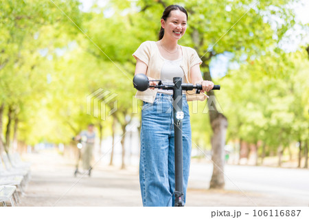 緑が美しい並木道を電動キックボードで爽快に走る若くてかわいい女性｜電動キックボードイメージ 106116887