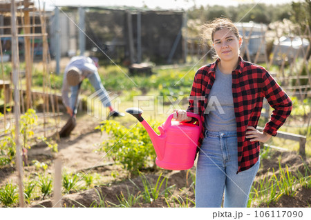 Skilled young woman farmer in plaid shirt holding pink watering can in hands and smiling at camera during work in vegetable garden in autumn 106117090