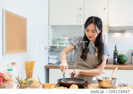 A beautiful young Asian girl in an apron is making pancakes in a minimalist home kitchen. A beautiful young Asian girl in an apron is making pancakes in a minimalist home kitchen. 106119897