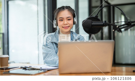 An attractive Asian female radio host is looking at her laptop screen, speaking into a microphone 106121886