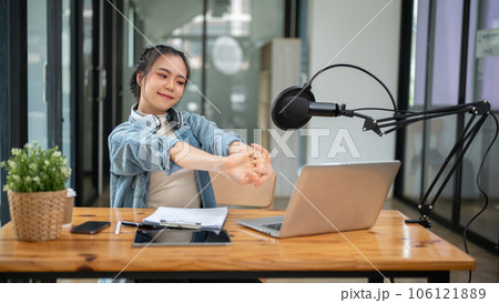 A happy Asian female radio host stretches her arms at her desk after finishing her live radio show A happy Asian female radio host stretches her arms at her desk after finishing her live radio show 106121889
