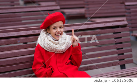 Smiling caucasian girl in a red coat and beret sits alone on a bench. Smiling caucasian girl in a red coat and beret sits alone on a bench. 106125006