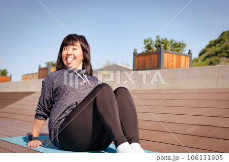 Sitting on a mat in a city park, a girl is doing yoga with a big smile on her face and her tongue hanging out. Concept: yoga, fitness, prank and fun 106131905