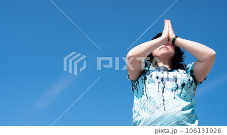 An impersonal portrait of a woman performing a set of yoga exercises, surya namaskara, holding her hands in front of her face against a blue sky 106131926