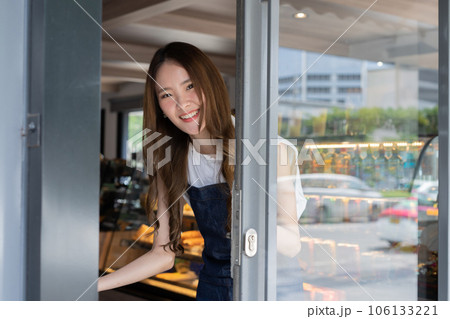 Young adult Asian female barista welcoming a customer walking into the coffee shop with a friendly smile and warm welcome. Small Business owner in Asia. Thai language sign on the door is to slide 106133221