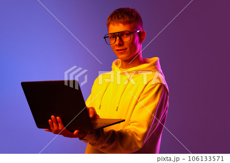 Young man, student in glasses standing with laptop against gradient purple background in neon light 106133571