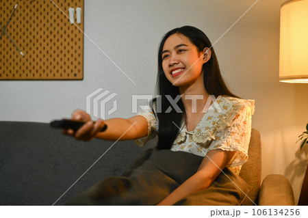 Happy asian woman watching television sitting on a comfortable sofa with the remote control in her hand 106134256