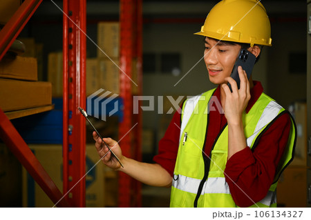 Storehouse male manager in reflective jacket talking on mobile phone while standing in a retail warehouse Storehouse male manager in reflective jacket talking on mobile phone while standing in a retail warehouse 106134327