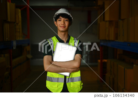 Confident male warehouse worker wearing safety vest holding laptop standing in large retail warehouse Confident male warehouse worker wearing safety vest holding laptop standing in large retail warehouse 106134340