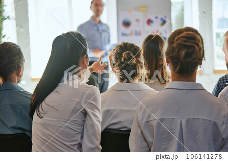 Rear view of woman pointing something in front of her while listening presentation against defocused leader, who teaching team of business people. Rear view of woman pointing something in front of her while listening presentation against defocused leader, who teaching team of business people. 106141278