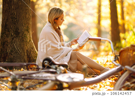 Beautiful young woman sitting on a fallen autumn leaves in a park, reading a book. Relaxation. 106142457