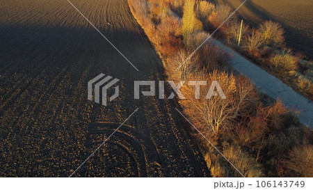 Beautiful landscape view old asphalt road with trees and shadows between large plowed agricultural fields of black soil on sunny autumn evening. Flying over dug agrarian fields of earth. Aerial drone Beautiful landscape view old asphalt road with trees and shadows between large plowed agricultural fields of black soil on sunny autumn evening. Flying over dug agrarian fields of earth. Aerial drone 106143749
