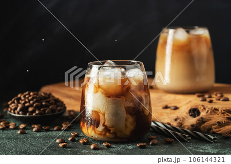Ice coffee in a tall glass with cream poured over, ice cubes and beans on a dark concrete table. 106144321