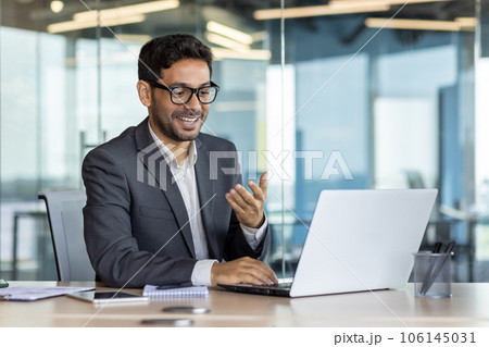 Young successful arab businessman inside office at workplace talking remotely with colleagues and partners, man using laptop for video call, satisfied boss smiling and gesturing with hands joyfully. 106145031