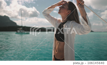 Woman standing on yacht deck looking at the horizon. Turquoise ocean water and mountains in background. Young long hair Caucasian female enjoys travel, tourism, holiday yacht tour to Bora Bora islands Woman standing on yacht deck looking at the horizon. Turquoise ocean water and mountains in background. Young long hair Caucasian female enjoys travel, tourism, holiday yacht tour to Bora Bora islands 106145640