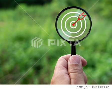 Human hand holding a magnifying glass with target icon on green nature background. Business concept. Management concept. Business planning concept. Human hand holding a magnifying glass with target icon on green nature background. Business concept. Management concept. Business planning concept. 106146432