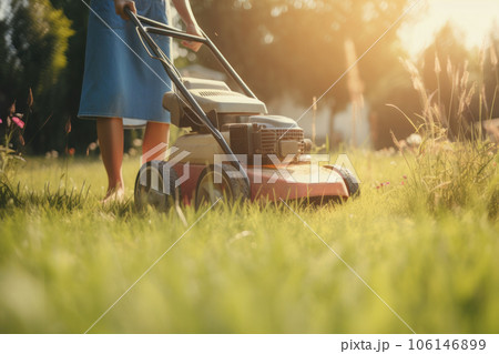 A woman in a hat pushing a manual lawnmower across a green yard on a sunny day is AI Generative. 106146899