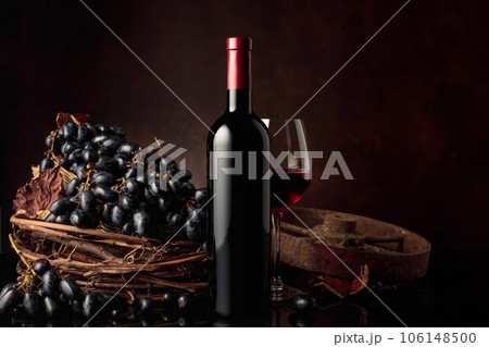 Glass and bottle of red wine with blue grapes on a black reflective background. 106148500