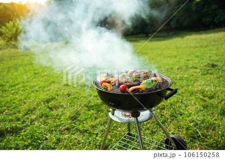 Grilled meat in grill. Roasting steaks and vegetables bbq in a round grill in a meadow. Summer picnic outdoors. Grilled meat in grill. Roasting steaks and vegetables bbq in a round grill in a meadow. Summer picnic outdoors. 106150258