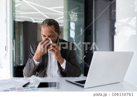 Sick man at workplace inside office with joy and cold, businessman sneezes sitting at desk, african american man with cold uses laptop at work. Sick man at workplace inside office with joy and cold, businessman sneezes sitting at desk, african american man with cold uses laptop at work. 106150761