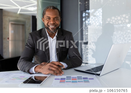 Portrait of mature successful adult African American businessman, boss smiling and looking at camera working inside office at workplace with laptop, satisfied investor financier behind paper work. Portrait of mature successful adult African American businessman, boss smiling and looking at camera working inside office at workplace with laptop, satisfied investor financier behind paper work. 106150765