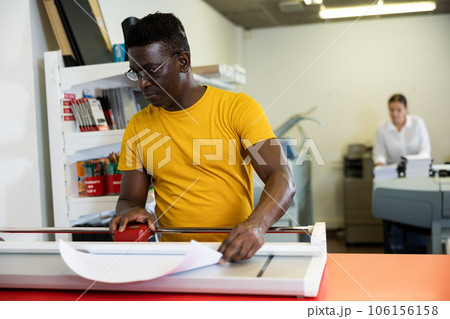 Focused middle-aged African American man cutting the printed sheet on the cutter in the typography Focused middle-aged African American man cutting the printed sheet on the cutter in the typography 106156158