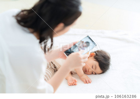リビングで昼寝をする赤ちゃんをスマホで撮影をする若いお母さん 106163286