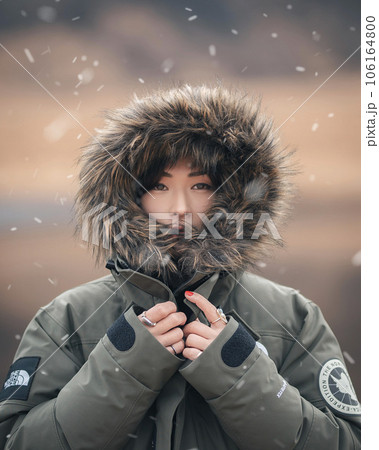 小雪舞う中ファーの付いたフードを被りこちらを見つめる女性 106164800