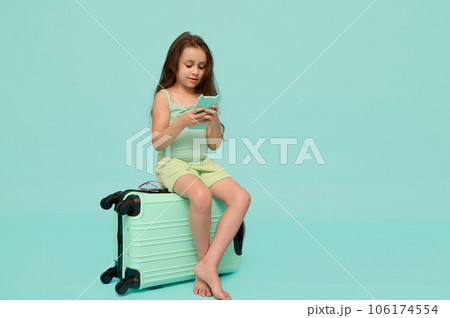 Little child girl using smartphone, sitting on suitcase, awaiting flight, checking mobile app, isolated blue background 106174554
