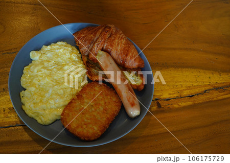 Breakfast consisting of hash browns, croissants, sausages, and scrambled eggs is served on gray ceramic plates placed on a wooden table. Breakfast consisting of hash browns, croissants, sausages, and scrambled eggs is served on gray ceramic plates placed on a wooden table. 106175729