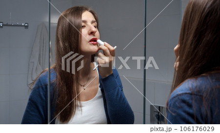 Portrait of young brunette woman looking in bathroom mirror and applying red lipstick. Self-made makeup, fashion and cosmetics Portrait of young brunette woman looking in bathroom mirror and applying red lipstick. Self-made makeup, fashion and cosmetics 106176029