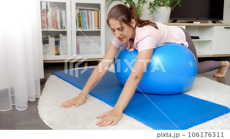 Happy smiling woman rocking and rolling on fitball during home fitness training. Concept of healthcare, sports and yoga at home. Happy smiling woman rocking and rolling on fitball during home fitness training. Concept of healthcare, sports and yoga at home. 106176311
