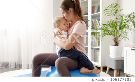 Young smiling woman holding her upset baby son and doing exercises on fitball at home. Concept of healthcare, sports and yoga at home Young smiling woman holding her upset baby son and doing exercises on fitball at home. Concept of healthcare, sports and yoga at home 106177185