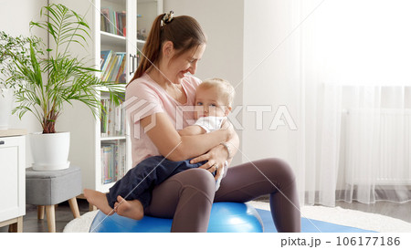 Young smiling woman doing fitness at home on fit ball while holding her little baby son Young smiling woman doing fitness at home on fit ball while holding her little baby son 106177186