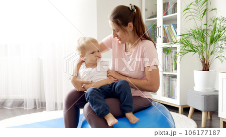 Young mother looking after her baby son while jumping and doing exercises on fitball in living room. Concept of healthcare, sports and yoga at home 106177187