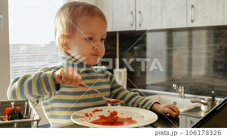 Cute messy boy eating soup with spoon and watching cartoons. Domestic food, gadget addiction, children healthy nutrition. Cute messy boy eating soup with spoon and watching cartoons. Domestic food, gadget addiction, children healthy nutrition. 106178326