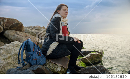 Young woman with long flowing hair sitting on a rocky coastline, holding a thermos of hot tea as she gazes out at the stormy ocean. Perfect for travel or holiday content. 106182630