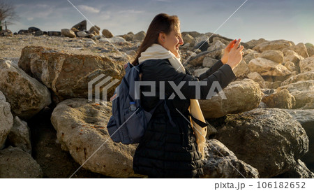 Young woman capturing the beauty of the winter sea by taking pictures with her smartphone while standing on the rocks. Ideal for vacation and travel. 106182652