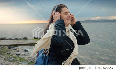 A smiling woman carrying a backpack walks along the beach while listening to music on a windy, chilly day. Essence of adventure, travel, and enjoyment. The perfect choice for travel-themed videos or A smiling woman carrying a backpack walks along the beach while listening to music on a windy, chilly day. Essence of adventure, travel, and enjoyment. The perfect choice for travel-themed videos or 106182723