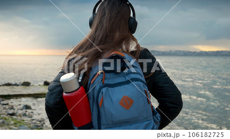 Smiling woman with a backpack, taking a walk on a cold, windy day on the beach while listening to music in headphones. It perfectly captures the essence of adventure, travel, and the joy of music, Smiling woman with a backpack, taking a walk on a cold, windy day on the beach while listening to music in headphones. It perfectly captures the essence of adventure, travel, and the joy of music, 106182725