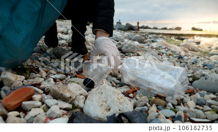 Young woman volunteer walking on the sea beach and collecting garbage and wastes in plastic bag. Concept of ecology, harm to environment, protecting nature, pollution and recycling problems. 106182735