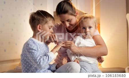 Smiling mother cutting fingernails to her two sons wearing pajamas at night. Parenting, child hygiene and healthcare, going to sleep. Smiling mother cutting fingernails to her two sons wearing pajamas at night. Parenting, child hygiene and healthcare, going to sleep. 106183680