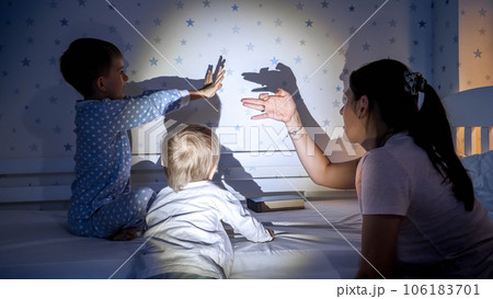 Cheerful kids with mother playing with shadows from the flashlight on wall at night. Family having time together, parenting, happy childhood and entertainment. 106183701