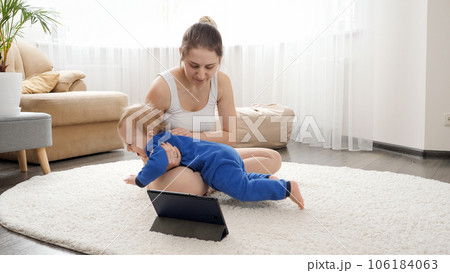 Young mother with baby boy watching online fitness course on tablet computer and doing exercises. Family healthcare, active lifestyle, parenting and child development Young mother with baby boy watching online fitness course on tablet computer and doing exercises. Family healthcare, active lifestyle, parenting and child development 106184063