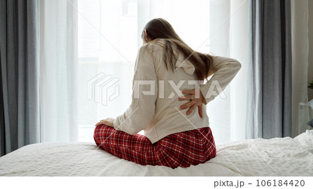 Young woman in pajamas sitting on the bed side and suffering from pain in back. Concept of problems with health, sickness, backache and pain. Young woman in pajamas sitting on the bed side and suffering from pain in back. Concept of problems with health, sickness, backache and pain. 106184420