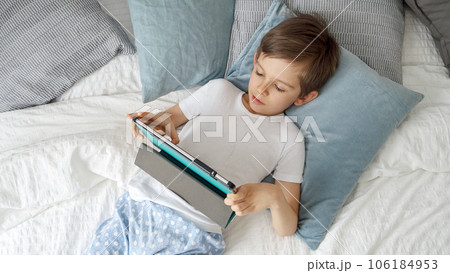 Little boy is lying on his bed, using a tablet computer during the day. Child using gadget, education and development. 106184953